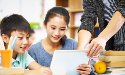 two asian elementary school children using digital tablet with help from teacher tutor in classroom.