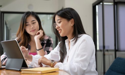 Horizontal photo of two female college student working on digital tablet and  talking about lessons comparing together.